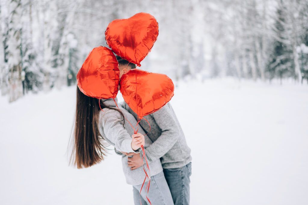 Loving couple cuddling in the winter forest holding heart-shaped balloons.