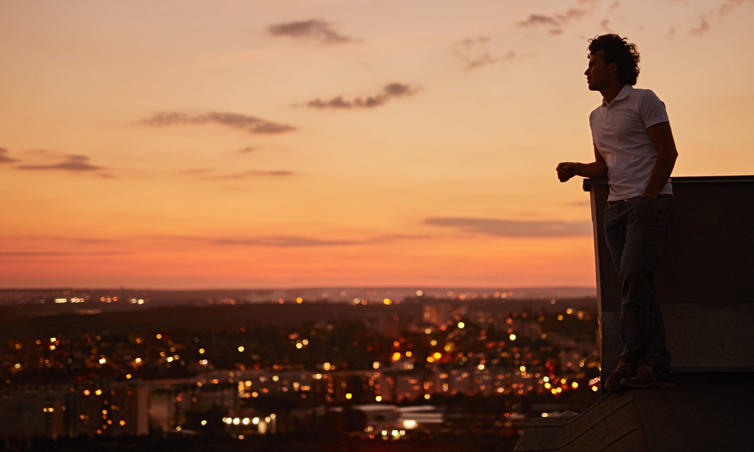 Low angle side view of unrecognizable tranquil contemplative male standing on rooftop terrace and admiring view of evening city at sunset time