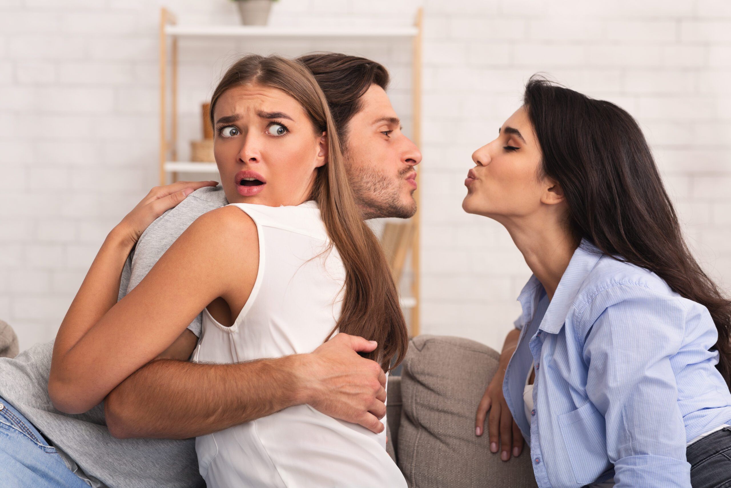 Love Triangle And Jealousy. Cheating Boyfriend Kissing A Girl While Hugging His Girlfriend Sitting Together On Couch Indoor.