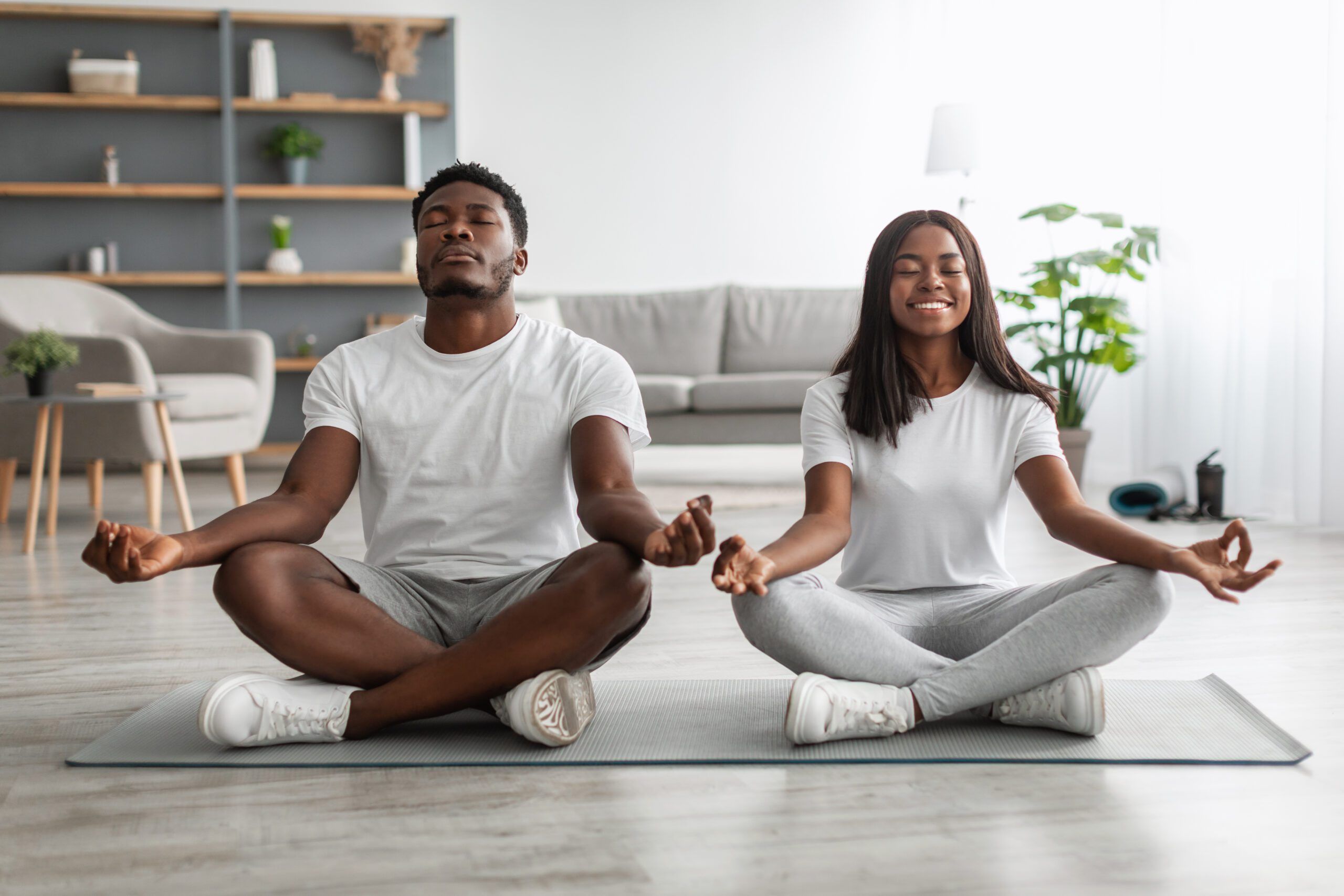 Domestic Yoga. Happy young black couple meditating with closed eyes at home in lotus position, millennial lady and guy sitting on fitness mat on the floor in living room, enjoying healthy lifestyle